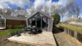 Modern lodge with large glass doors, a wooden deck, outdoor seating, and trees in the background under clouds.