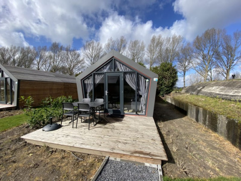 Cabaña moderna con grandes ventanales, terraza de madera y sillas exteriores, rodeada de árboles y cielo nublado.