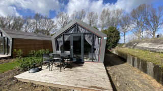 Cabaña moderna con grandes ventanales, terraza de madera y sillas exteriores, rodeada de árboles y cielo nublado.