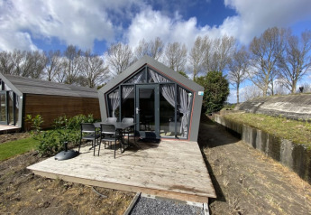 Moderne lodge met glazen deuren, houten terras en tuinstoelen, omgeven door bomen en bewolkte lucht.