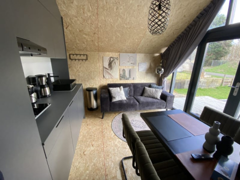 Interior de lodge moderno con cocina, sofá gris, mesa de comedor y ventanas grandes hacia el jardín.