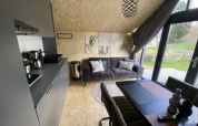 Interior de lodge moderno con cocina, sofá gris, mesa de comedor y ventanas grandes hacia el jardín.