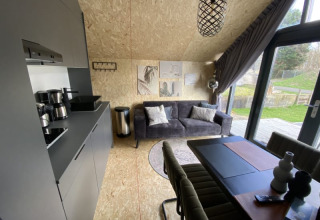 Interior de lodge moderno con cocina, sofá gris, mesa de comedor y ventanas grandes hacia el jardín.
