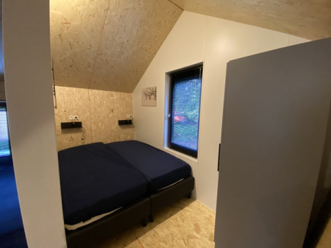 Soveværelse i en lodge med skrå loft, to senge med mørkt sengetøj, vindue og lyst træpanel.