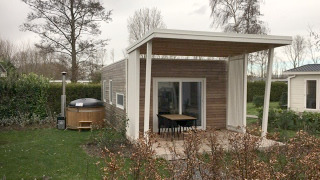 Modern tiny house with a covered patio and outdoor hot tub in a green garden with trees and bushes.