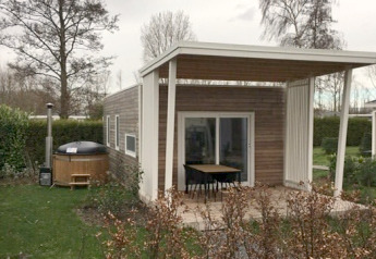 Modern tiny house with a covered patio and outdoor hot tub in a green garden with trees and bushes.