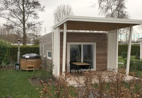 Modern tiny house met overdekt terras en een buiten hottub in een groene tuin met bomen en struiken.
