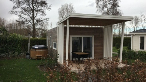 Modern tiny house with a covered patio and outdoor hot tub in a green garden with trees and bushes.