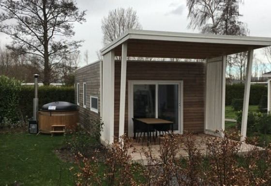 Modern tiny house with a covered patio and outdoor hot tub in a green garden with trees and bushes.