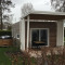 Modern tiny house met overdekt terras en een buiten hottub in een groene tuin met bomen en struiken.