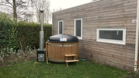 Klein houten huisje met twee ramen en een ronde buitenspa op het gras naast het huis.