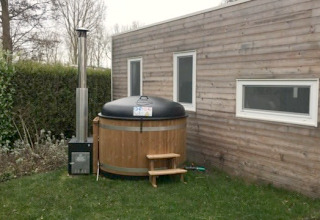 Klein houten huisje met twee ramen en een ronde buitenspa op het gras naast het huis.