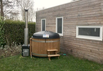 Tiny wooden house with two windows and a round outdoor hot tub on the lawn next to the house.