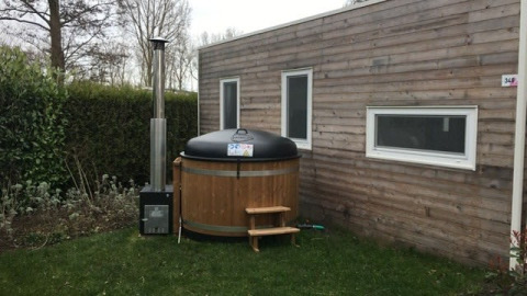 Tiny wooden house with two windows and a round outdoor hot tub on the lawn next to the house.