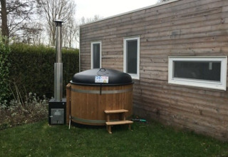 Tiny wooden house with two windows and a round outdoor hot tub on the lawn next to the house.