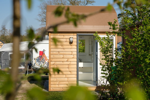 Buitenfoto van een moderne glampingcabine met houten gevel, open deur en fietsen op de achtergrond.