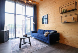 Moderne glampingaccommodatie met een blauwe zetel, houten wand, grote ramen en knusse decoratie.