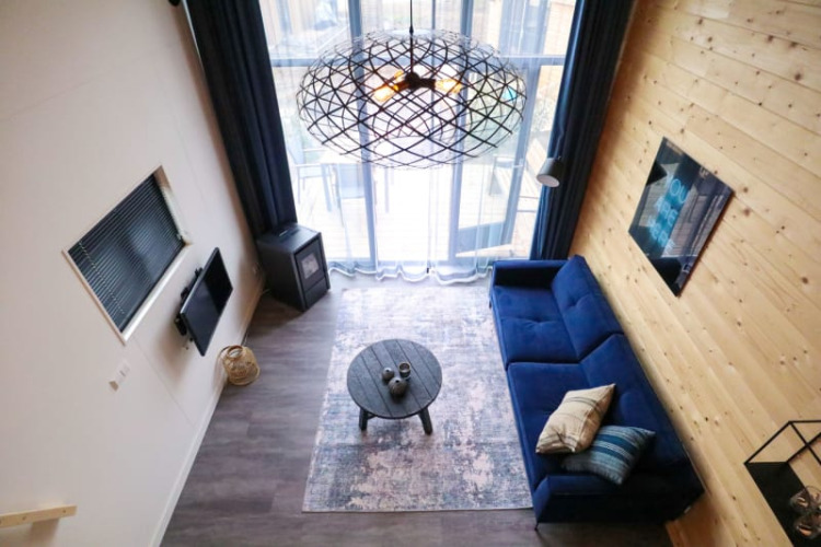 Alojamiento glamping moderno con sofá azul, mesa redonda y ventanales grandes vistos desde arriba.