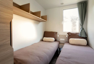 Lichte lodgekamer met twee aparte bedden, bruine lakens, houten rekken en raam met een gordijn.