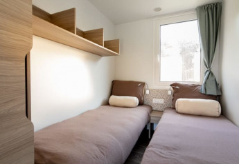 Chambre lumineuse en lodge avec deux lits simples, literie marron, étagères en bois et fenêtre avec rideau.