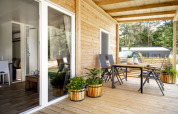 Terrasse en bois d’un lodge moderne avec table, chaises et plantes en pot, entourée de forêt paisible.