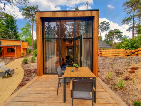 Ein kleines Holzhaus mit großen Fenstern und Terrasse mit Sitzbereich, umgeben von Wald und Natur.