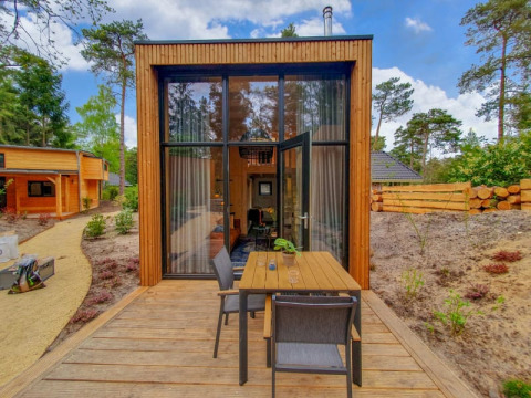Pequeña casa de madera con grandes ventanales y terraza al aire libre, rodeada de árboles y naturaleza.