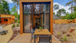 Klein houten huis met grote ramen en buitenterras, gelegen in een bosrijke omgeving met veel groen.