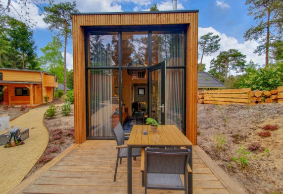 Pequeña casa de madera con grandes ventanales y terraza al aire libre, rodeada de árboles y naturaleza.