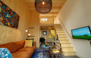 Modern tiny house living room with wooden walls, loft area, brown sofa, colorful art, and TV.