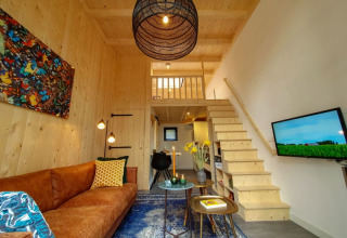 Modern tiny house living room with wooden walls, loft area, brown sofa, colorful art, and TV.