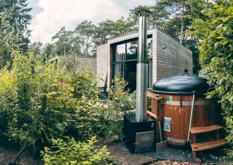 Photo of a modern glamping cabin in the forest with a wood-fired hot tub surrounded by lush greenery.