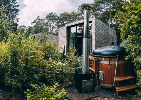 Photo of a modern glamping cabin in the forest with a wood-fired hot tub surrounded by lush greenery.