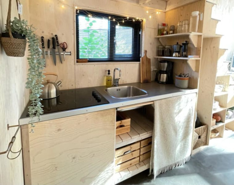 Petite cuisine d'une tiny house avec bois clair, évier, étagères ouvertes et plaque de cuisson électrique.