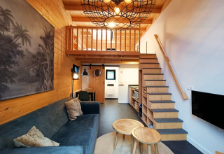 Modernes Tiny House Wohnzimmer mit Hochbett, Holzdekor, Sofa, Couchtischen und Treppe zum Schlafbereich.