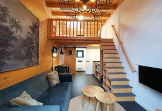 Modern tiny house living room with loft, wooden interior, blue sofa, round tables, and staircase to bed.