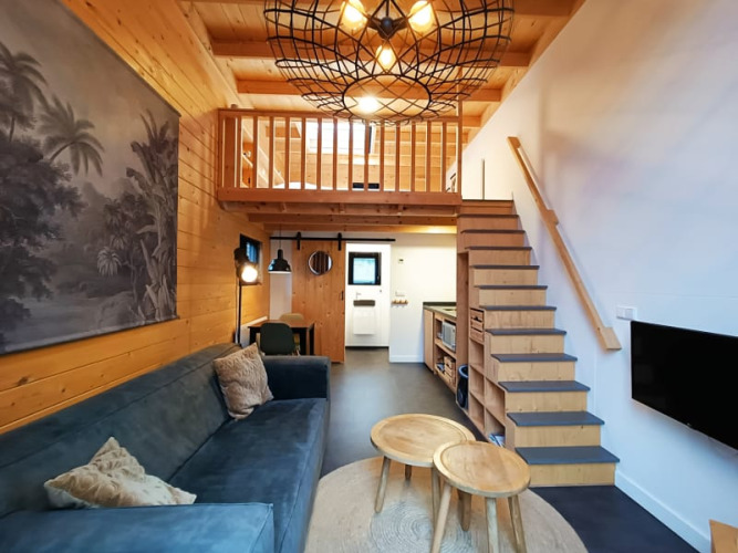 Salon de tiny house moderne avec mezzanine, intérieur bois, canapé bleu et escalier menant à la chambre.