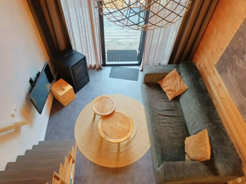 Woonkamer van een tiny house van bovenaf met bank, ronde tafels, tv, houtkachel en terrasdeur.