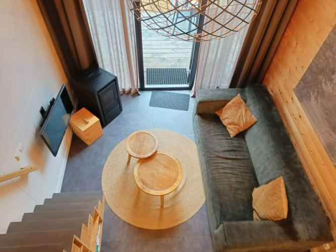 Salon d'une tiny house vu d'en haut, avec canapé, tables rondes, télévision et grande porte-fenêtre.