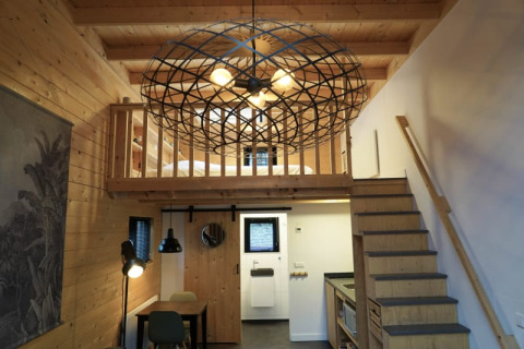 Modern tiny house interior with wooden loft, staircase, kitchen, desk, and a large geometric chandelier.