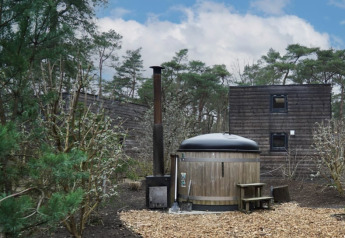 Petite maison en bois avec sauna extérieur rond, entourée de nature et d’arbres sous un ciel partiellement bleu.