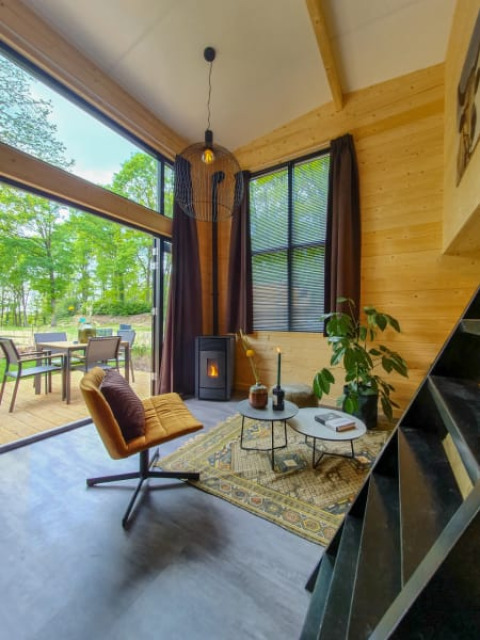 Soggiorno accogliente in tiny house con pareti in legno, grandi finestre e vista su terrazza esterna.