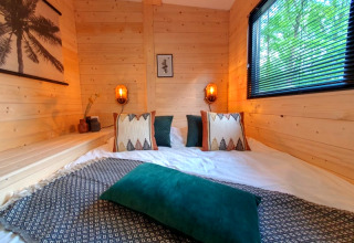 Cozy bedroom in a tiny house, featuring wood-paneled walls, patterned pillows, a window, and green scenery outside.