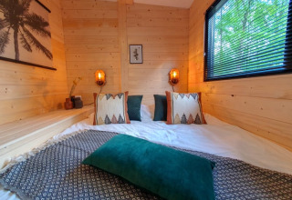 Cozy bedroom in a tiny house, featuring wood-paneled walls, patterned pillows, a window, and green scenery outside.
