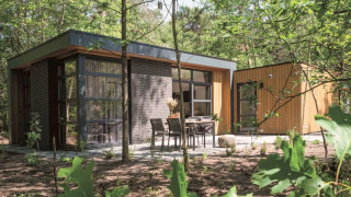 Lodge moderne entouré de forêt, grandes baies vitrées et terrasse extérieure aménagée avec table et chaises.