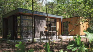 Lodge moderne entouré de forêt, grandes baies vitrées et terrasse extérieure aménagée avec table et chaises.