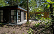 Modern lodge with large windows and flat roof, nestled in a wooded area surrounded by green trees.