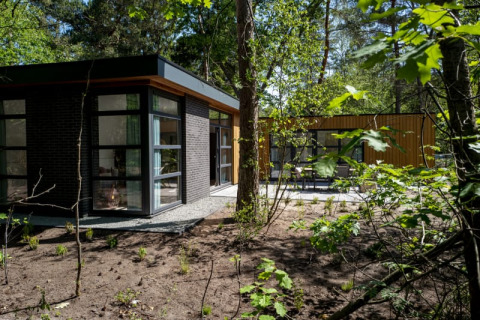 Modern lodge with large windows and flat roof, nestled in a wooded area surrounded by green trees.