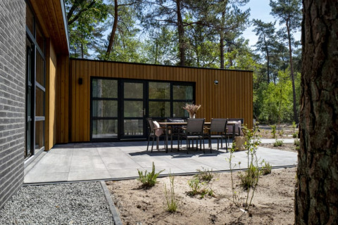 Lodge moderne à façade en bois, grande terrasse pavée et salon de jardin, au cœur d'une forêt verdoyante.