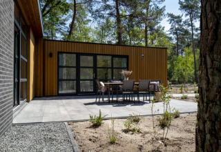 Lodge moderne à façade en bois, grande terrasse pavée et salon de jardin, au cœur d'une forêt verdoyante.